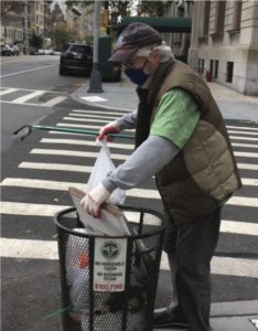 Mark Smith in green T-shirt and brown puffer vest and baseball cap in the street, putting trash into a wire trash can.