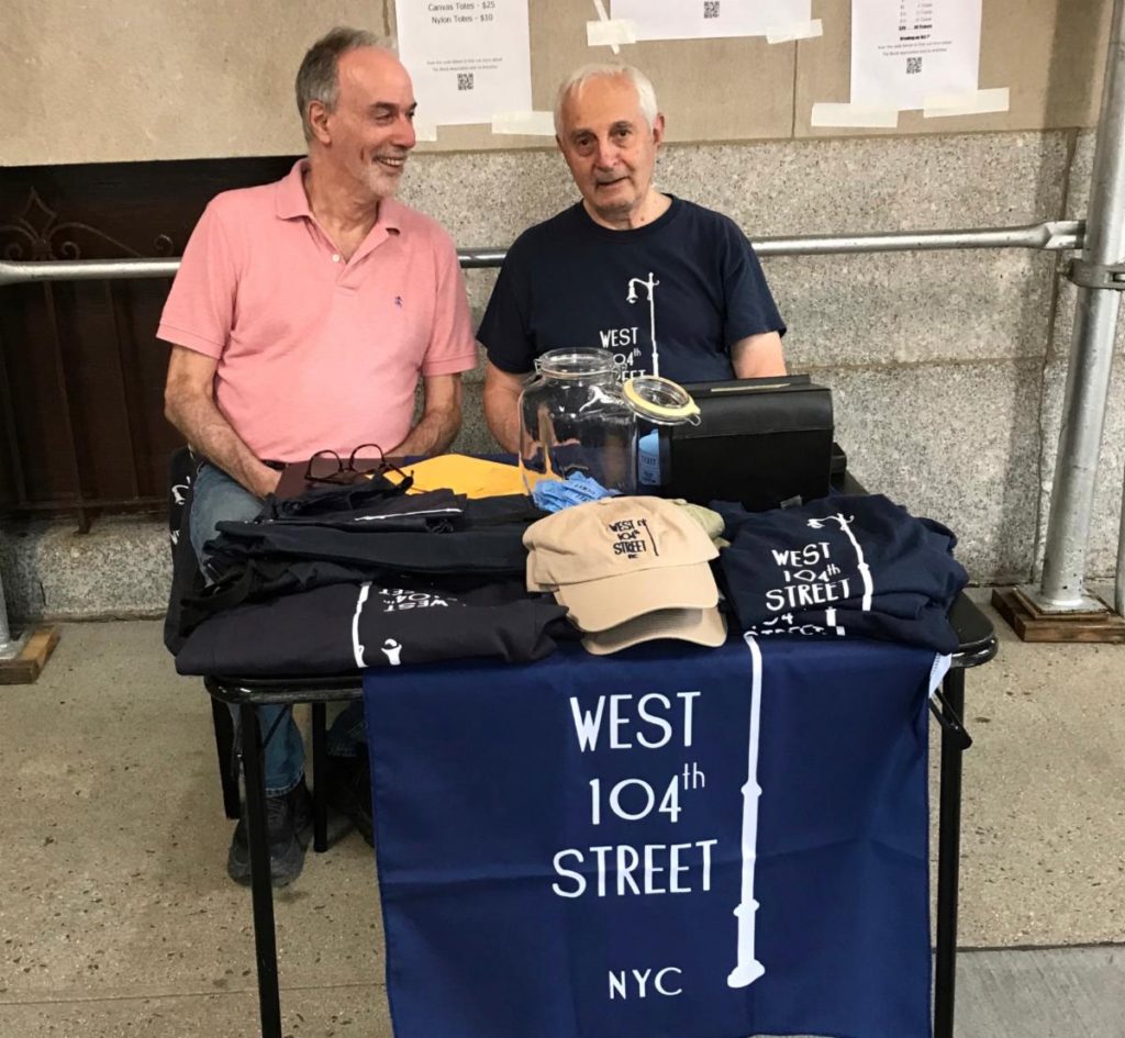 Two men, block residents, sitting at a table selling hats, t-shirts and raffle tickets for the West 104th Street Block Association yard sale, October 1.