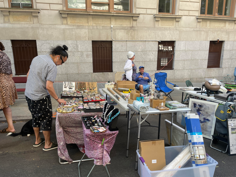 Woman browsing a jewelry table at the street fair as people pass by and man observes from a set of blue lawn chairs.