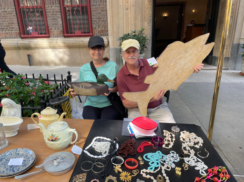 A woman holding a wooden duck decoy and a man holding a large, wooden carving of a fish sitting at a table with tableware and costume jewelry.