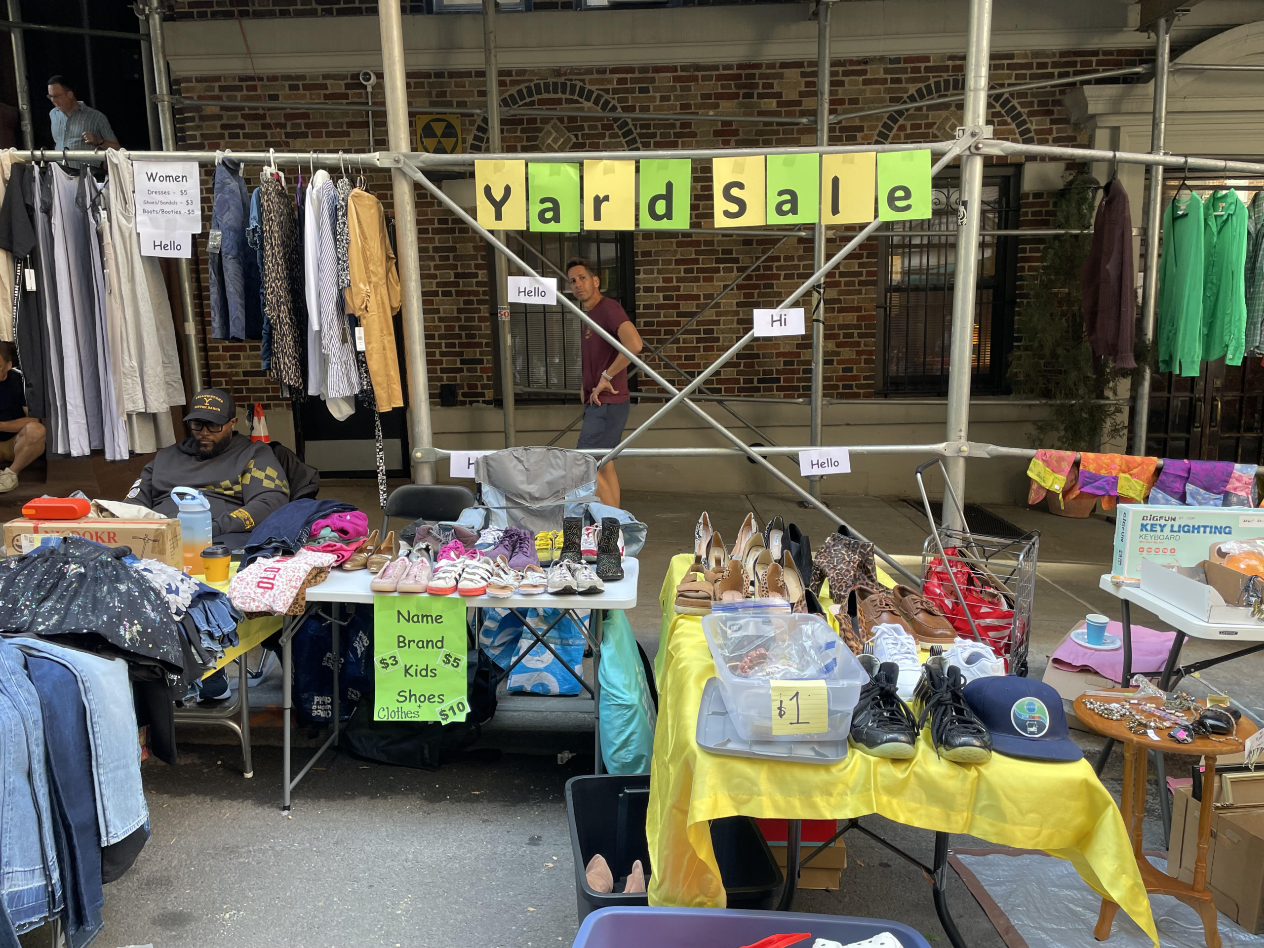 Street scene with tables of shoes and blue jeans and a yellow and green “Yard Sale” banner and clothing items hanging from building scaffolding.
