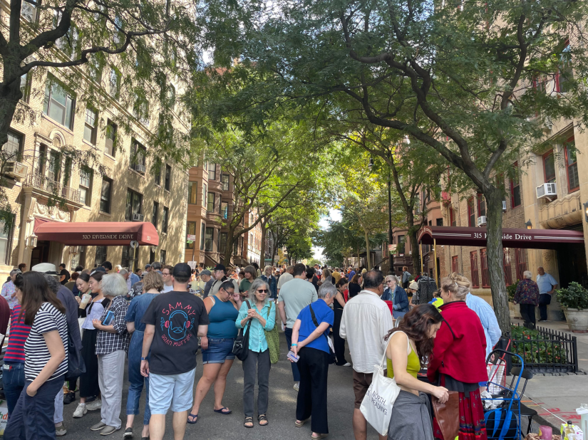 Crowd milling about west 104th street between residential buildings with brown awnings and arcing trees