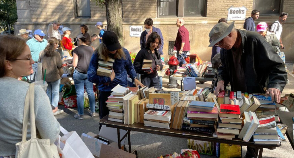 Street fair book sale table full of books with several people browsing. Two women have several books in their arms. A man in glasses and a fishing hat browses titles. A woman in glasses in the foreground looks to the side, holding two light colored totes.