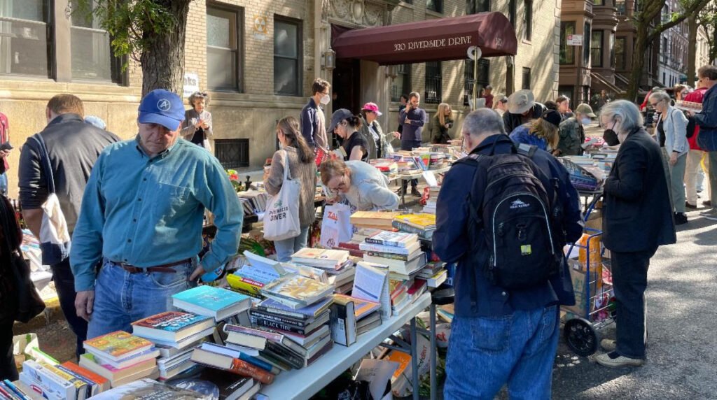 Street view of book sale table at the West 104th Street Block Association yard sale. Several people holding totes and wearing backpacks or pushing carts are browsing. The burgundy awning from 320 Riverside Drive is visible in the background along with a few brownstones and trees.