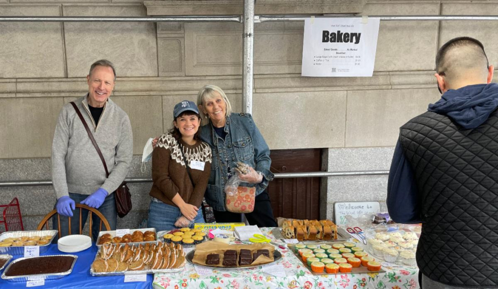 A man in blue latex gloves and two women smile in front of a table filled with cookies, brownies and cupcakes.
