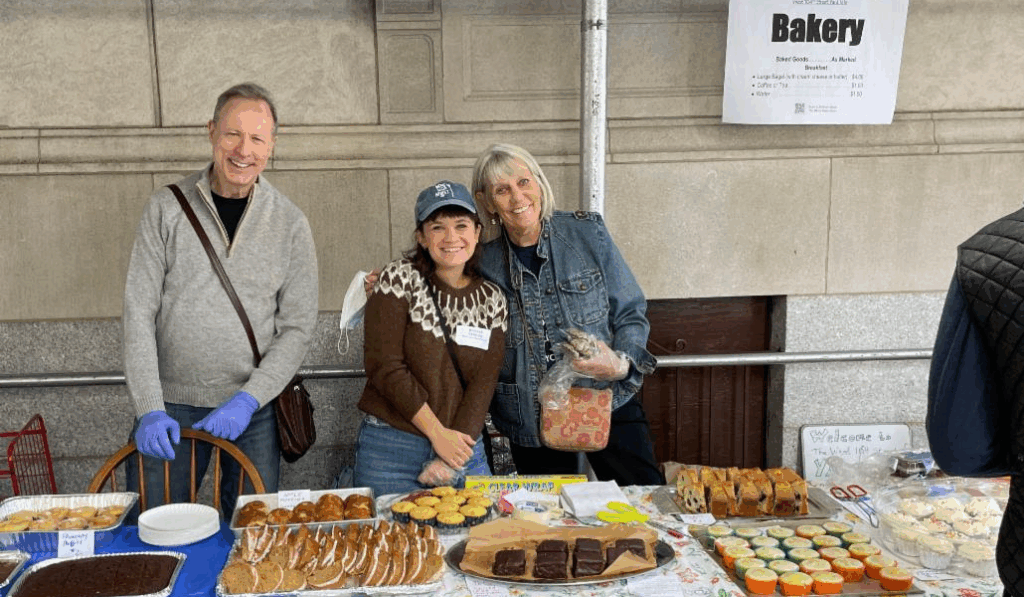 A man in blue latex gloves and two women smile in front of a table filled with cookies, brownies and cupcakes.