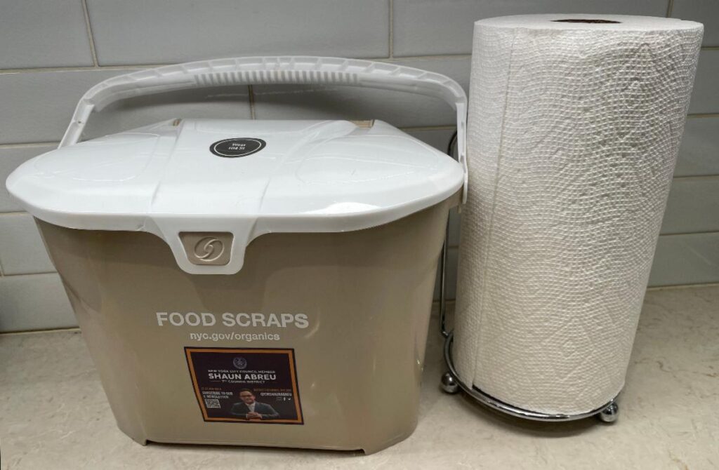 Tan colored Composting pail with a white handle and lid shown on a kitchen counter next to a roll of paper towels.