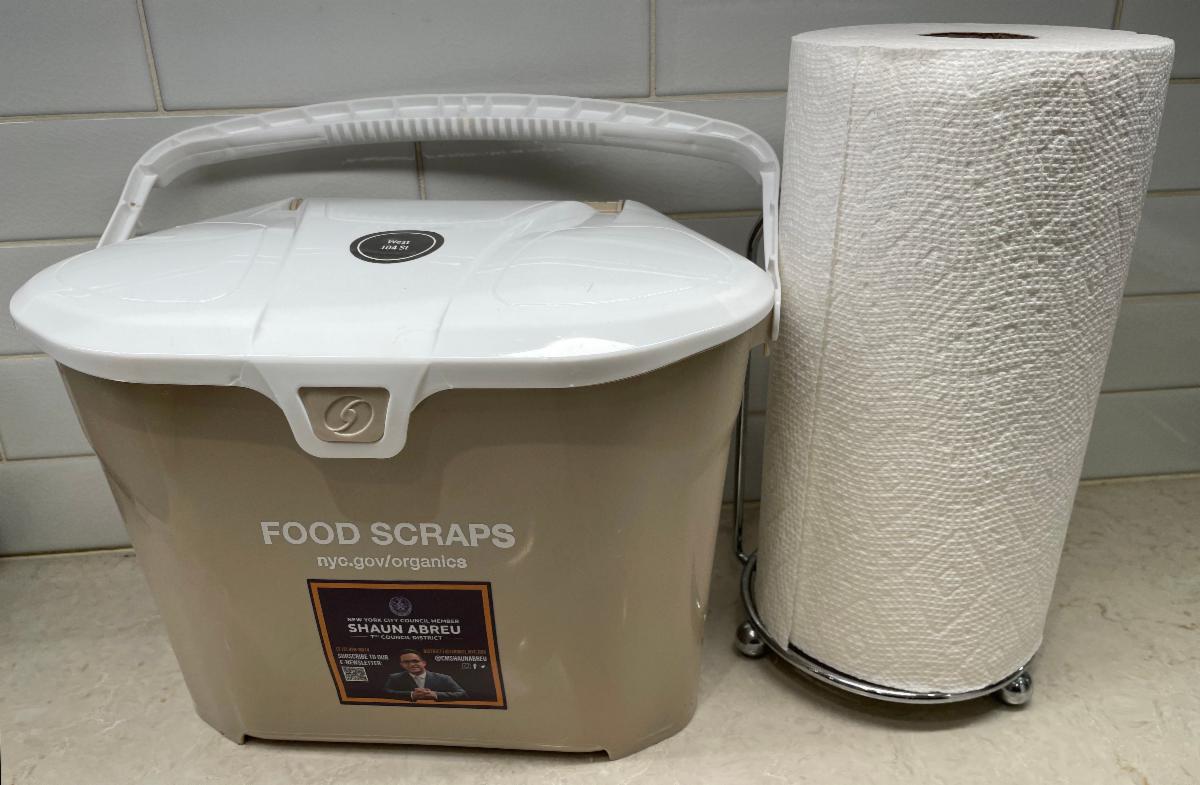 Tan colored Composting pail with a white handle and lid shown on a kitchen counter next to a roll of paper towels.