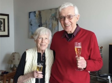 Photograph of Eloy and Bob Ledogar holding glasses of champagne in their living room. Elderly white couple. Woman with white hair, black elbow length blouse and white ves sanding next to a taller elderly white haired man in glasses and a red sweater one a black collared shirt. Both are smiling and holding a glass of champagne.