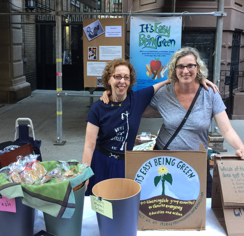 Two smiling women wearing glasses and tshirts stand behind a sidewalk table. A sign on the table and a banner behind them say “It’s Easy Being Green. Three buckets on the table labeled trash and recycle . The one in the middle is full of cellophane wrapped baked goods set in a green plaid kitchen towel.
