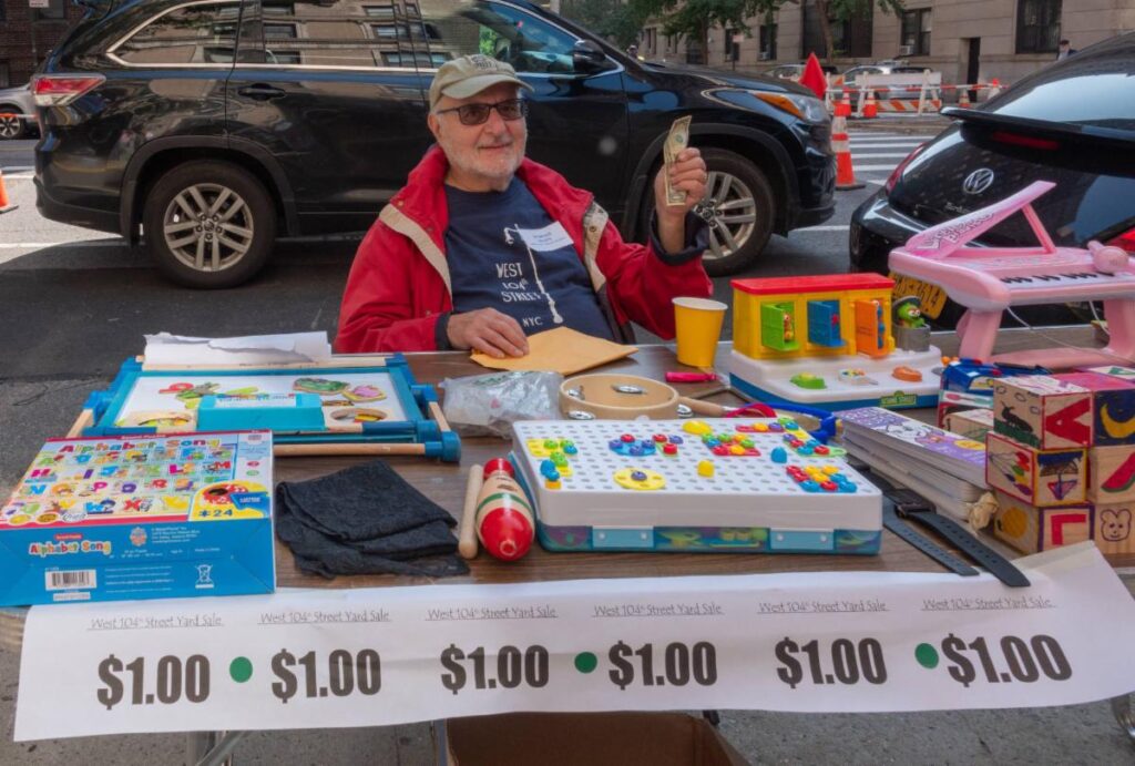 Smiling man in a red jacket and tan cap sitting at a table full of bargain items with a banner in front and a parked car behind him.