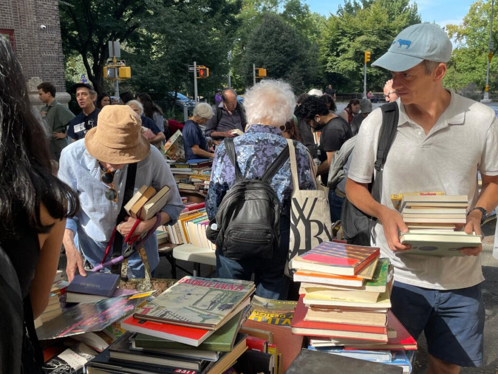 Street scene. Stacks of books on a table with a person in a tan bucket hat bent over the table holding three books, a woman with short, white hair and a black backpack facing away in center and a man in a white polo and light blue baseball cap caring a stack of books at the right.