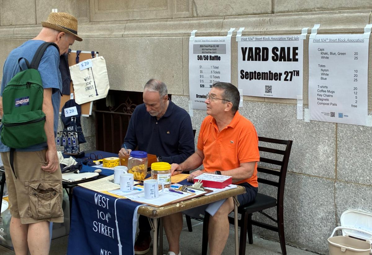 A man in a dark shirt and a man in an orange polo sitting at a table with a sign on the building wall behind them with the words, “YARD SALE September 27th”. Another man wearing a green backpack, a straw hat, blue tshirt and tan shorts stands over the table looking at the two men.