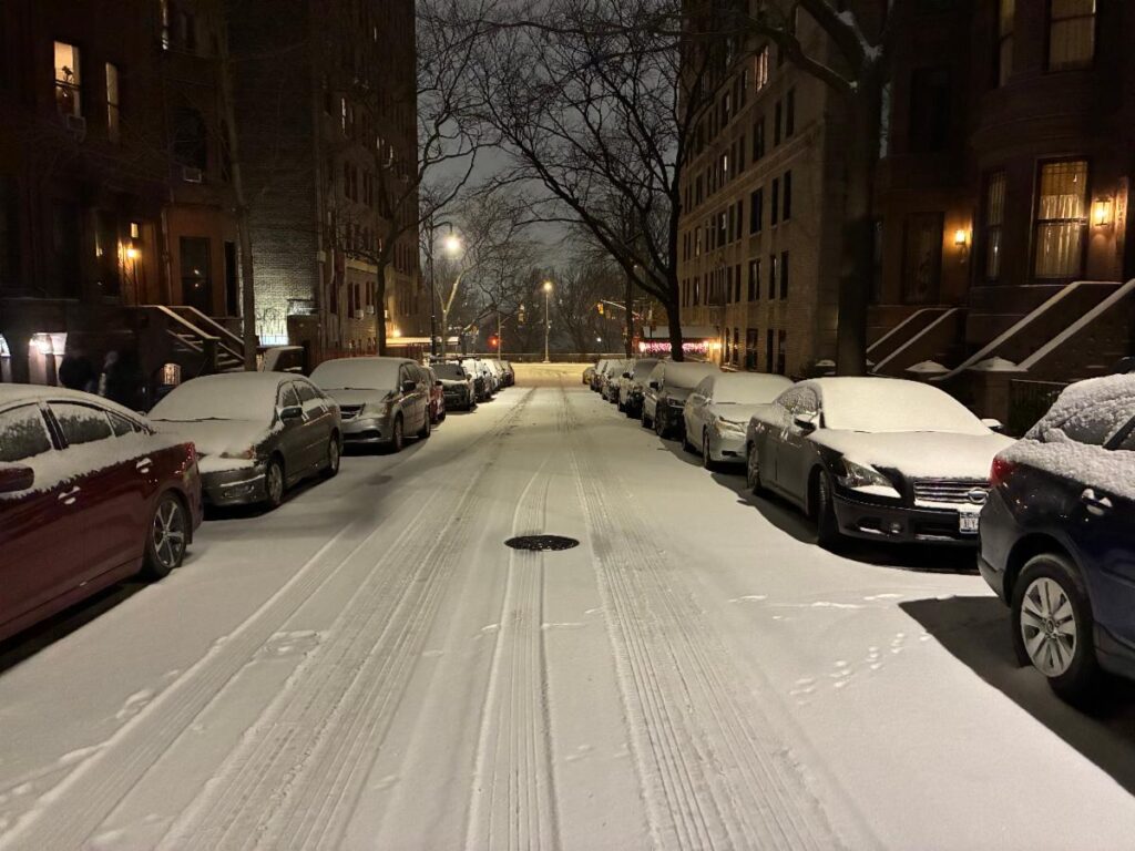 View at night down a snowy street with snow-covered cars and tall, brick buildings lining both sides. A tree lit up by a street light in the distance.