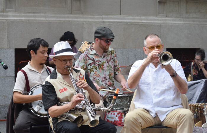 Group of four seated men playing banjo, drums and alto saxophone and trumpet