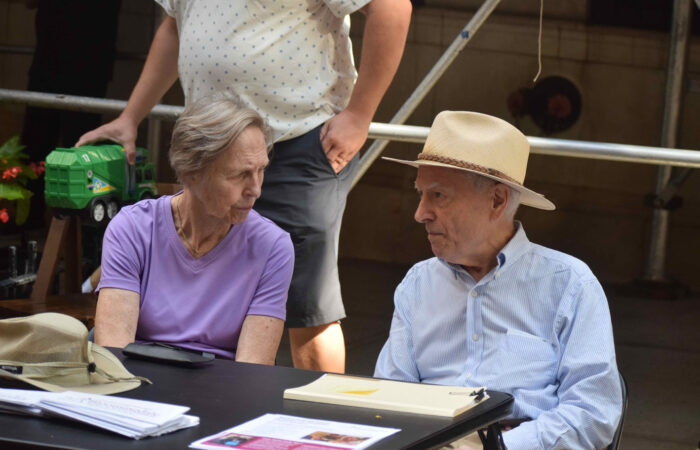 An elderly woman in with short gray hair and a lavender t-shirt is seated at a table next to an elderly man wearing a blue futtondown and a straw hat. A woman in a white tshirt stands benind them.