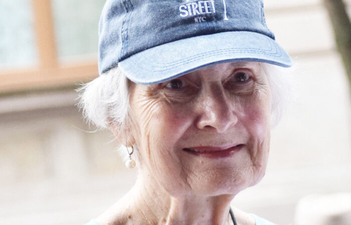 An elderly woman with short gray hair smiles while wearing a light blue West 104th Street Block Association baseball cap
