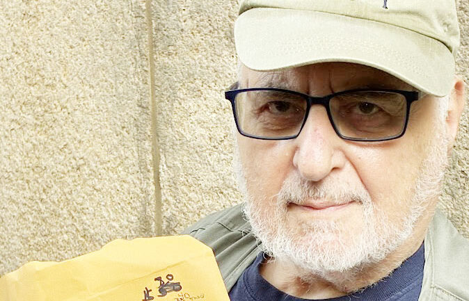 An elderly man with a tan West 104 block association baseball cap and black glasses holding a manila envelope