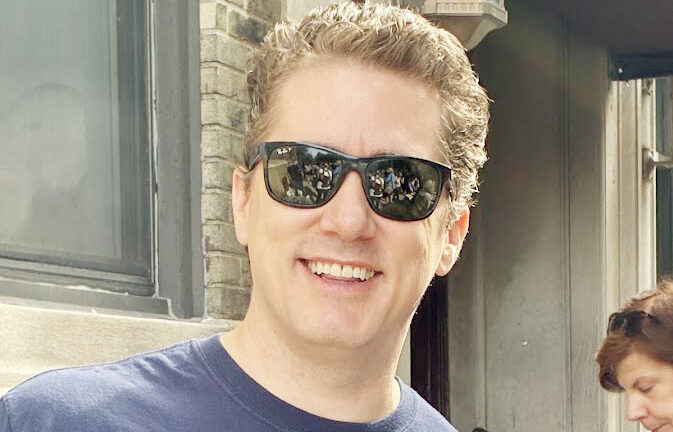 A blond man in dark sunglasses and a blue-gray West 104th Street t-shirt