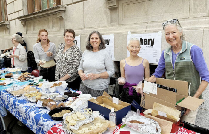 Six women smiling behind a table full of baked goods. The farthest of the six is helping a customer.