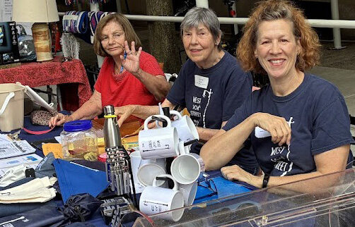 Three seated women and a stack of white coffee mugs, seated behind a talbe with blue and red cloths and a raffle tub