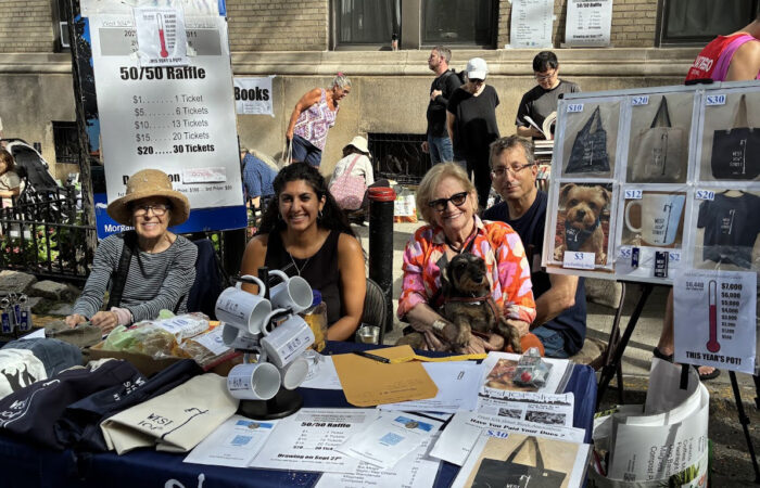 Three women and a man sit behind the silent auction table with large posters of raffle items and shoppers browsing on the sidewalk behind them.