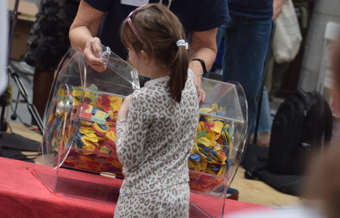A small girl, back turned, wearing a ponytail, white and brown-spotted leapard print outfit reaches into the raffle bin as a red-haired woman in a navy t-shirt looks on