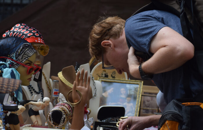 A person with short red hair wearing a navy t-shirt and glasses, bent over a table full of jewelry, silk scarves and gloves.