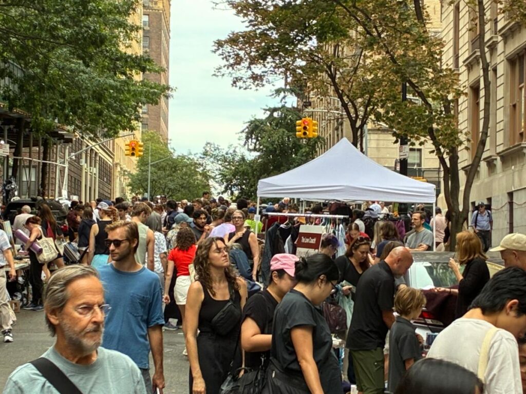 Yard sale street scene view looking west toward Riverside Park. People walking along the street center. A white tent canopy and traffic lights are visible around tables of goods for sale along West 104th Street.