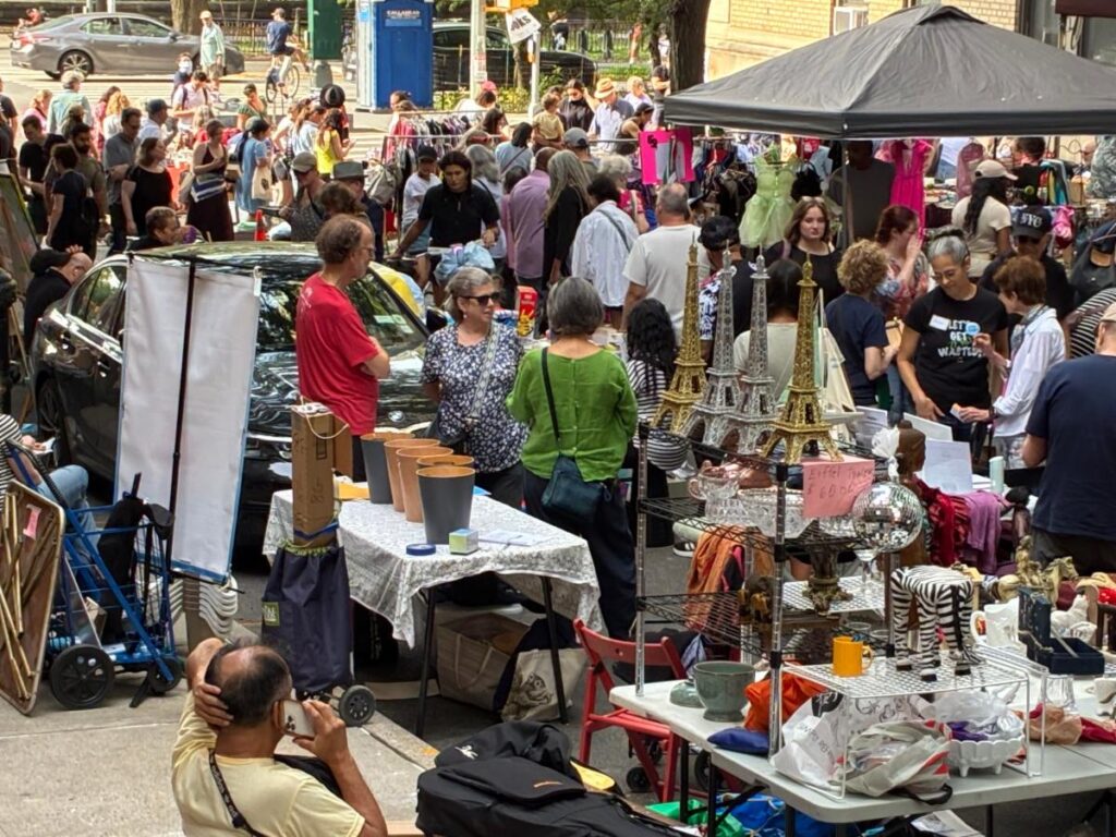 Yard sale street scene along West 104th Street. People gathering around tables of goods for sale, including two gold and two silver, large models of the Eiffel Tower for sale