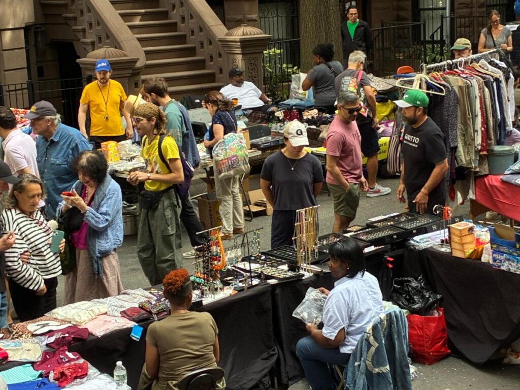 Yard sale street scene along West 104th Street, taken from above. People gathering around tables of clothing and jewelry for sale,