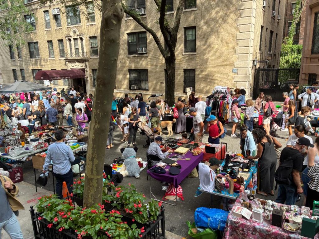 Yard sale street scene. People gathering around tables of goods for sale along West 104th Street.