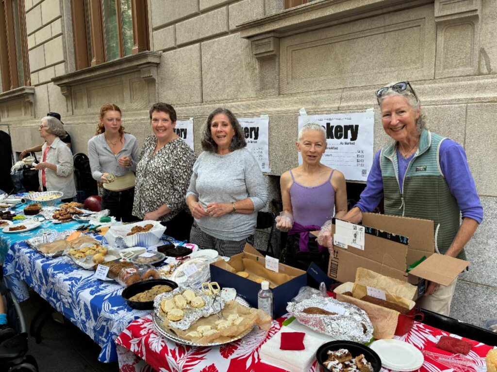 Bake Sale 2025 a line of smiling women standing behindrr a long table fof homemade baked goods with blue and white & red and white tropical print tablecloth