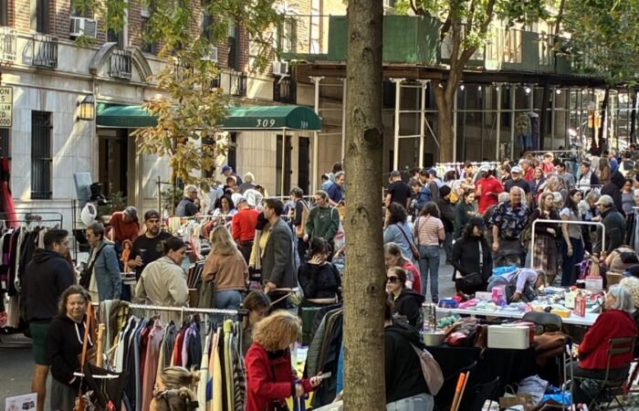 A street scene showing yard sale shoppers in front of the green awning of 309 W. 104th St. A narrow true trunk divides the photo in half.