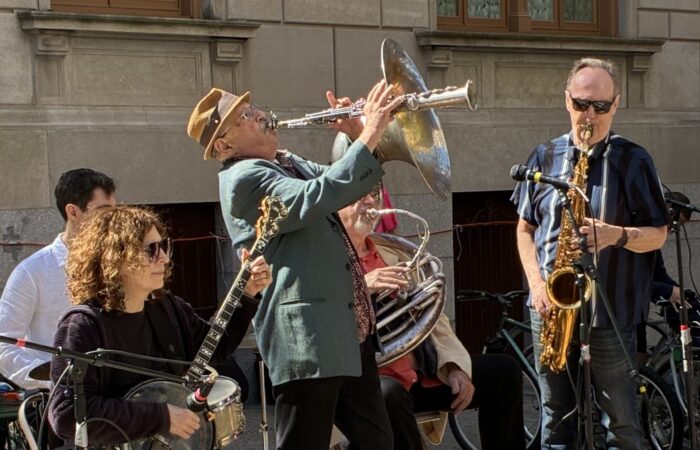 Five jazz band members, including a drummer and tuba player in the back of obscured by a seated female guitarist wearing sunglasses, a trumpet player in a green jacket and pork pie hat holding his horn up high and a saxophone player in sunglasses