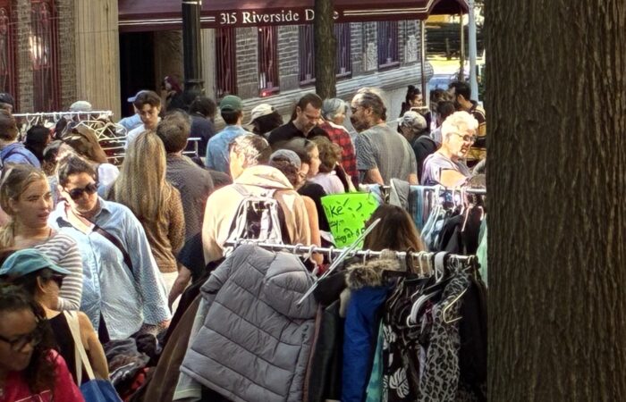Another street scene featuring yard sale shoppers among racks of clothing. Tree bark frames the right side of the photo.
