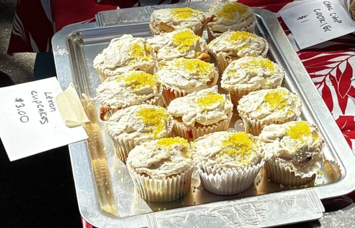 A tray of lemon cupcakes with white frosting sprinkled with lemon zest on a red and white tropical print tablecloth. A sign says “lemon cupcakes $3.00”